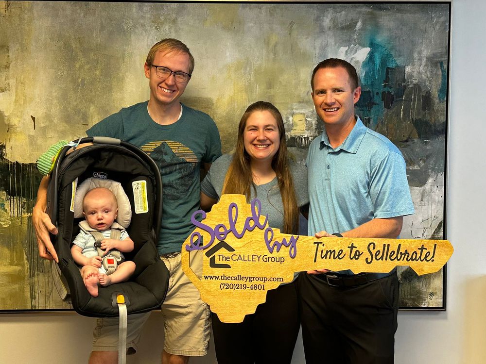 Steve Calley with a young couple and their baby at closing, holding his key-shaped Time to Sellebrate sign.