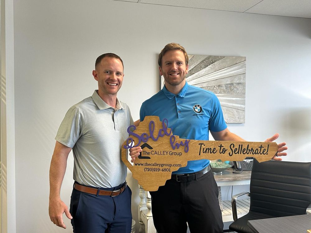 Steve Calley with a client in a blue polo holding the Time to Sellebrate key sign in an office.