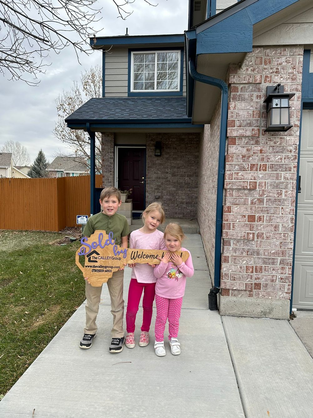 Three young children holding the Welcome Home key sign on the front walk of their family's new home.