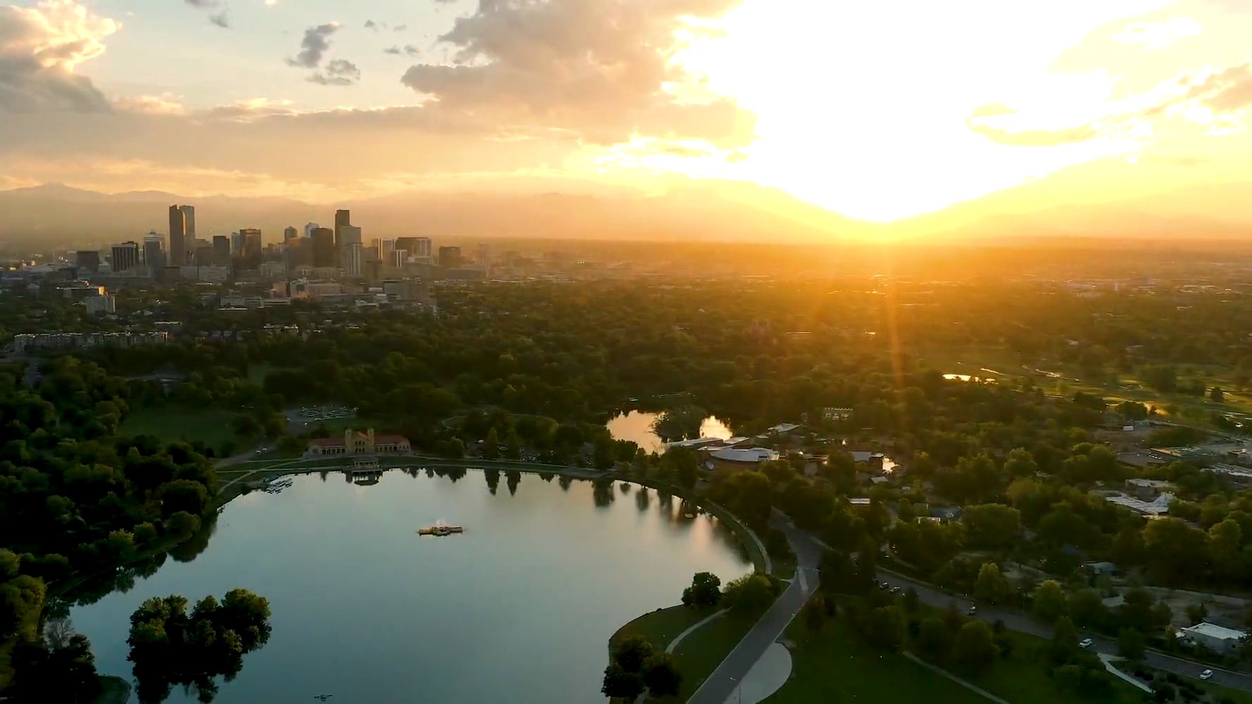 Aerial view of Denver's City Park and downtown skyline at sunset, with the Front Range beyond.