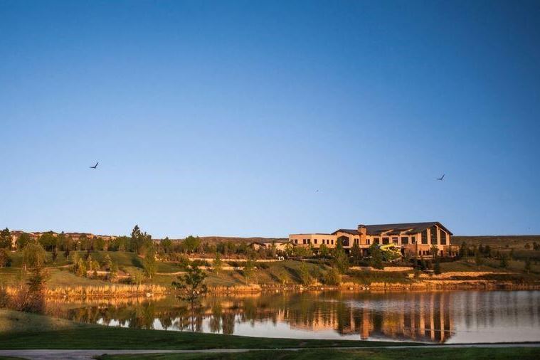 A still pond reflecting a lodge-style clubhouse on the rise beyond it, lit gold by the evening sun, with two birds in a clear sky.