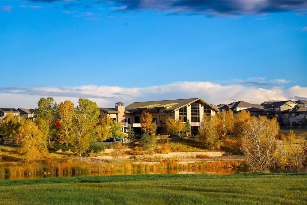 A lodge-style clubhouse with tall windows above a pond, ringed by yellow and orange autumn trees under a deep blue sky.