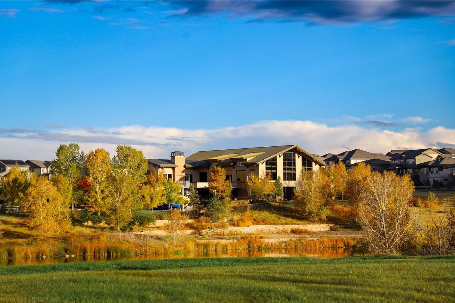 A lodge-style clubhouse with tall windows above a pond, ringed by yellow and orange autumn trees under a deep blue sky.