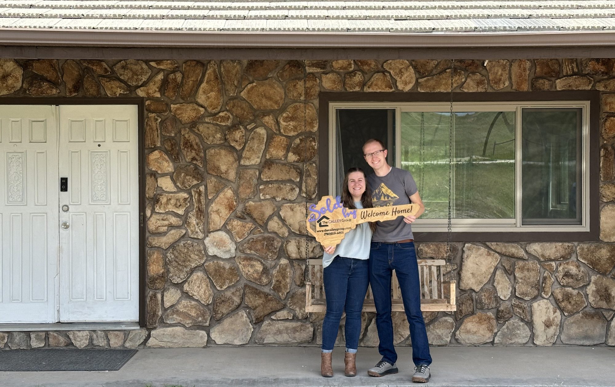 A couple standing at the front door of their stone-front home, holding Steve Calley's key-shaped closing sign.